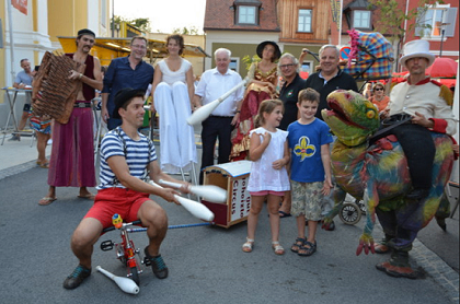Eine Gruppe von Menschen steht auf einer Straße. Ein Mann jongliert mit Stöcken, und ein Mädchen in einem weißen Shirt steht neben ihm. Hinter ihnen sind ein Dinosaurierkostüm und ein Kinderwagen zu sehen. Menschen stehen hinter ihnen, und Gebäude sind im Hintergrund.