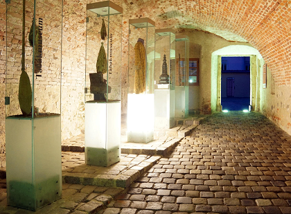 Ein schwach beleuchteter Flur mit Ziegelwänden, der mehrere Glasvitrinen mit Skulpturen und Artefakten zeigt. Der Flur führt zu einer beleuchteten Tür.