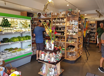 Ein Mann in einem blauen Shirt und Shorts ordnet Produkte auf einem Regal in einem Geschäft an. Der Laden hat Holzregale voller verschiedener Artikel, eine Kühlvitrine mit Obst und Gemüse und Menschen beim Einkaufen.