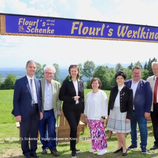 Eine Gruppe von Personen steht vor einem Schild mit der Aufschrift Flourl's Schenke. Sie sind geschäftlich gekleidet. Hinter ihnen steht eine Holzbank. Der Hintergrund zeigt ein Grasfeld mit Bäumen und Bergen.