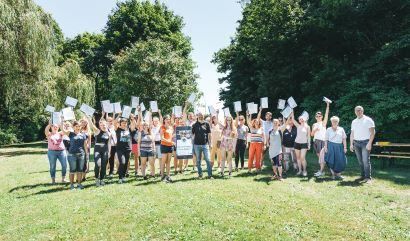 Eine Gruppe von Menschen steht auf einem Rasen und hält weiße Papiere in der Hand. Sie posieren für ein Foto. Im Hintergrund sind Bäume und eine Bank zu sehen.