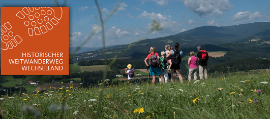 Eine Gruppe von Wanderern steht auf einem Grashügel und betrachtet die Berglandschaft. Sie tragen Rucksäcke und Hüte, einer von ihnen trägt einen gelben Hut.