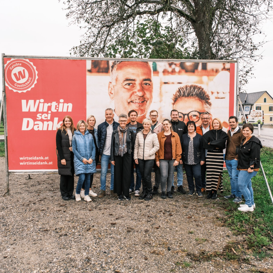Eine Gruppe von Menschen posiert für ein Foto vor einem großen roten Schild mit der Aufschrift 'Wirtin sei Dank'. Das Schild zeigt das Gesicht eines lächelnden Mannes. Sie sind in Winterkleidung gekleidet.