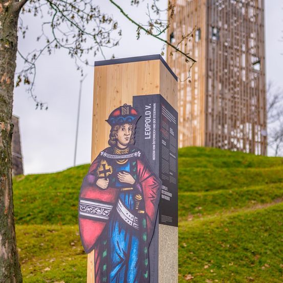 Ein hölzerner Pfosten mit einer gemalten Figur eines mittelalterlichen Ritters steht auf einem Grashügel. Der Ritter hält ein Kreuz und ein Schwert und trägt einen roten Umhang und einen blauen Mantel. Dahinter steht ein hoher Turm.