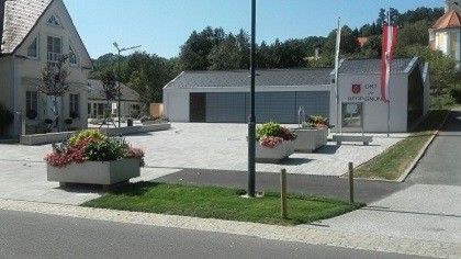 Ein kleines Dorf mit einem großen weißen Gebäude in der Mitte. Das Gebäude hat zwei Flaggen, eine rote und eine weiße. Es gibt zwei Blumenbeete auf beiden Seiten des Gebäudes.