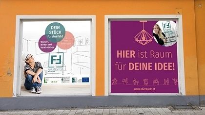 Ein orangefarbenes Gebäude hat zwei Fenster mit Postern, die einen Arbeitsbereich bewerben. Eine Frau sitzt auf dem Boden in der Nähe des Fensters. Das Poster sagt: 'Hier ist Raum für deine Idee!'