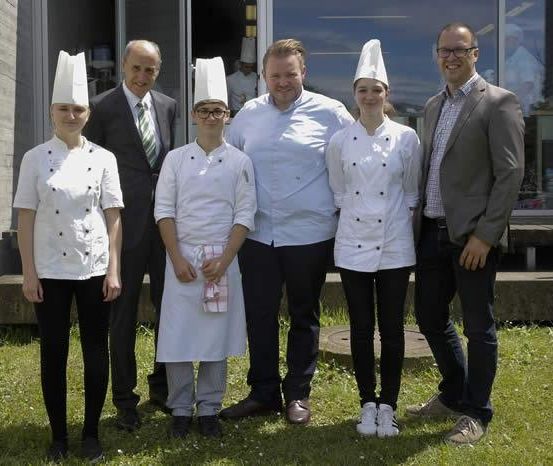 Fünf Personen stehen vor einem Gebäude mit einer Glaswand. Sie tragen alle Kochmützen und weiße Uniformen. Sie lächeln und posieren für ein Foto. Dahinter ist eine verschwommene Ansicht einer Küche mit einer Person in einer Kochmütze.