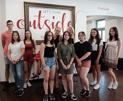 Eine Gruppe junger Menschen posiert für ein Foto vor einem Schild mit der Aufschrift 'Let's Think Outside'. Das Schild hat einen Bilderrahmen dahinter. Sie tragen Freizeitkleidung.
