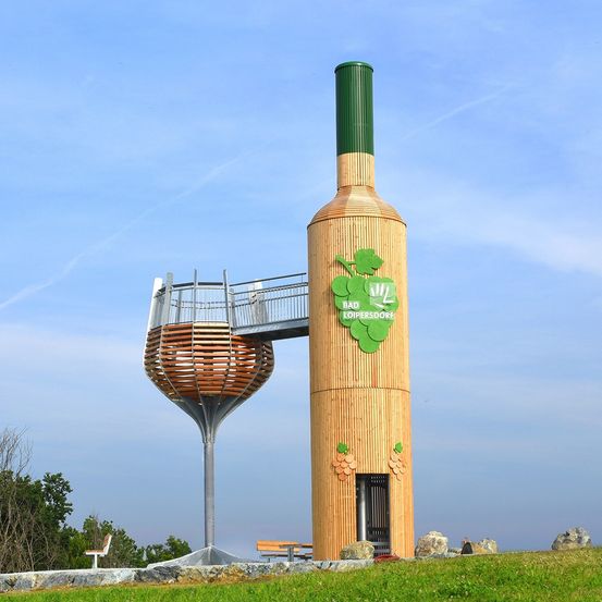 Eine hölzerne Weinflaschenskulptur mit einem befestigten Weinkelch steht auf einem Hügel. Sie hat einen Balkon auf dem Weinkelch und ein grünes Logo mit Text an der Seite.