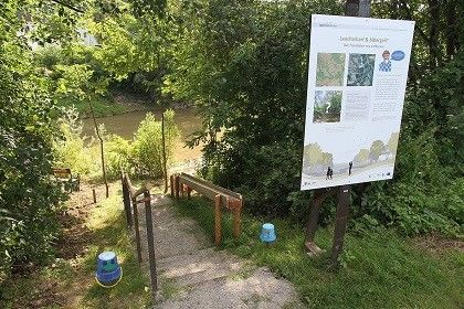 Ein Schild mit Informationen steht in der Nähe eines Flusses, mit einer Holzbank und Stufen, die zum Wasser führen. Bäume und Grün umgeben das Gebiet. Ein kleiner blauer Behälter ist in der Nähe platziert.