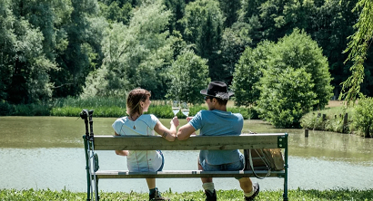 Ein Paar sitzt auf einer Bank am See, Weinflaschen in der Hand, lächelt und genießt die Aussicht.