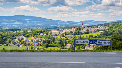 Ein Straßenschild zeigt die Richtungen nach Graz und Wien an. Die Straße ist leer, und im Hintergrund befindet sich ein Dorf mit einem Turm, umgeben von grünen Hügeln und einem klaren Himmel.