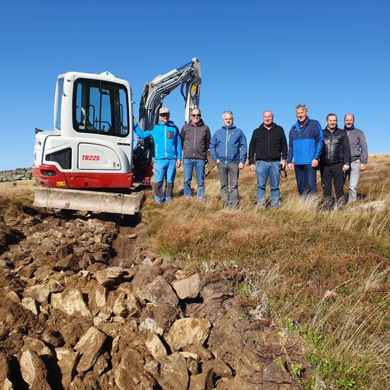 Sechs Männer stehen auf einem Grashügel, mit einem TB225-Bagger hinter ihnen. Die Männer sind in Jacken und Hosen gekleidet. Der Bagger ist rot und weiß und hat eine Schaufel daran. Die Männer lächeln und scheinen für ein Foto zu posieren.