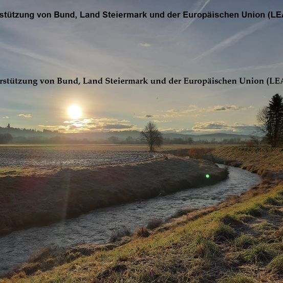 Eine Landschaft mit einem kleinen Fluss und einem untergehenden Sonnen. Die Szene zeigt ein flaches, trockenes Feld, spärliche Bäume und Berge in der Ferne. Ein Wasserzeichen lautet 'Unterstützung von Bund, Land Steiermark und der Europäischen Union (LEA)'.