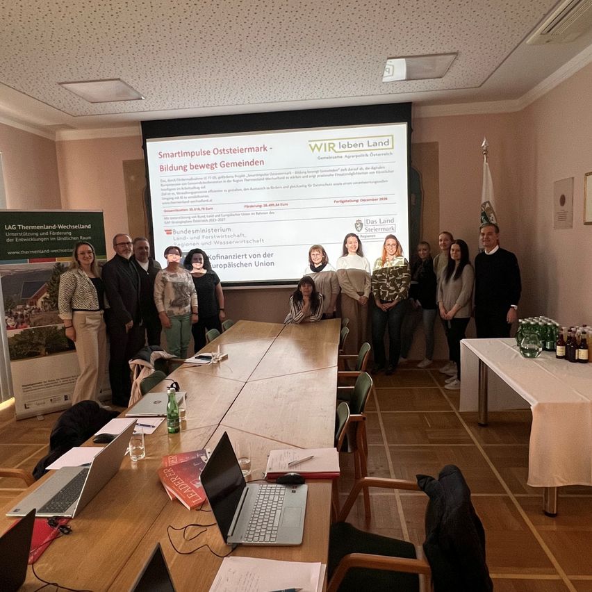 Eine Gruppe von Menschen steht in einem Konferenzraum mit Laptops und Papieren. Eine Projektionsleinwand zeigt eine Präsentation über 'Smartimpulse Osteiermark - Aufbau besserer Gemeinden'. Ein Tisch hat Flaschen, Gläser und eine Flagge.