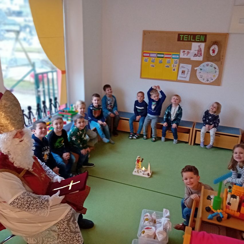 Ein Mann, der als Sankt Nikolaus verkleidet ist, liest ein Buch vor einer Gruppe von Kindern, die auf einer Bank und einem Stuhl in einem Raum sitzen.