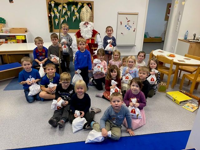 Eine Gruppe von Kindern und ein Erwachsener, der als Weihnachtsmann verkleidet ist, sitzen auf dem Boden in einem Klassenzimmer. Sie alle halten Tüten, möglicherweise Geschenke.