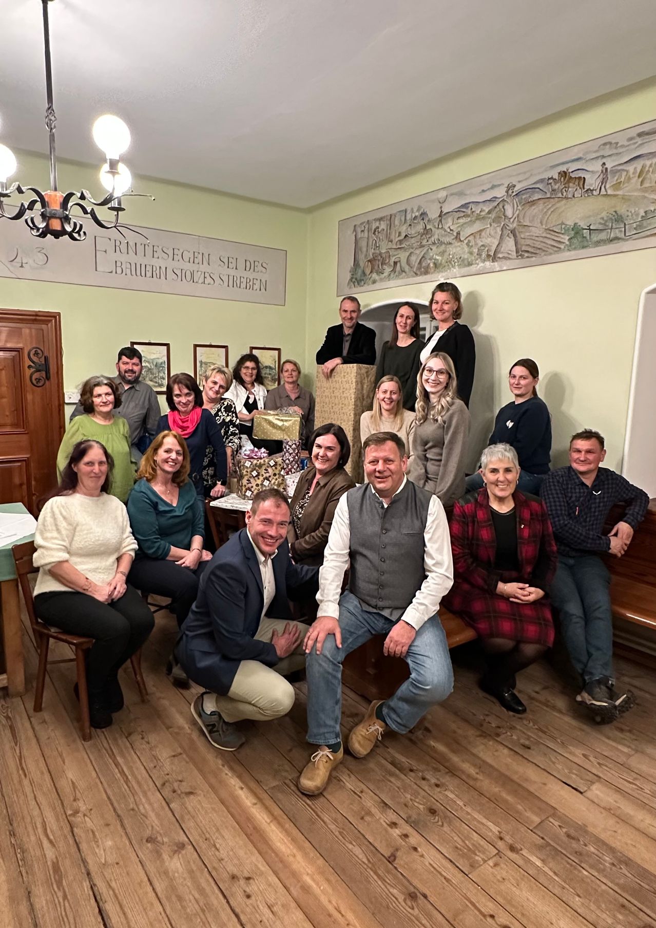 Eine Gruppe von Menschen, sowohl Männer als auch Frauen, posiert für ein Foto in einem Raum. Sie sitzen auf einer Bank und Stühlen, mit ein paar Geschenken auf dem Tisch. Dahinter hängt ein Banner an der Wand.