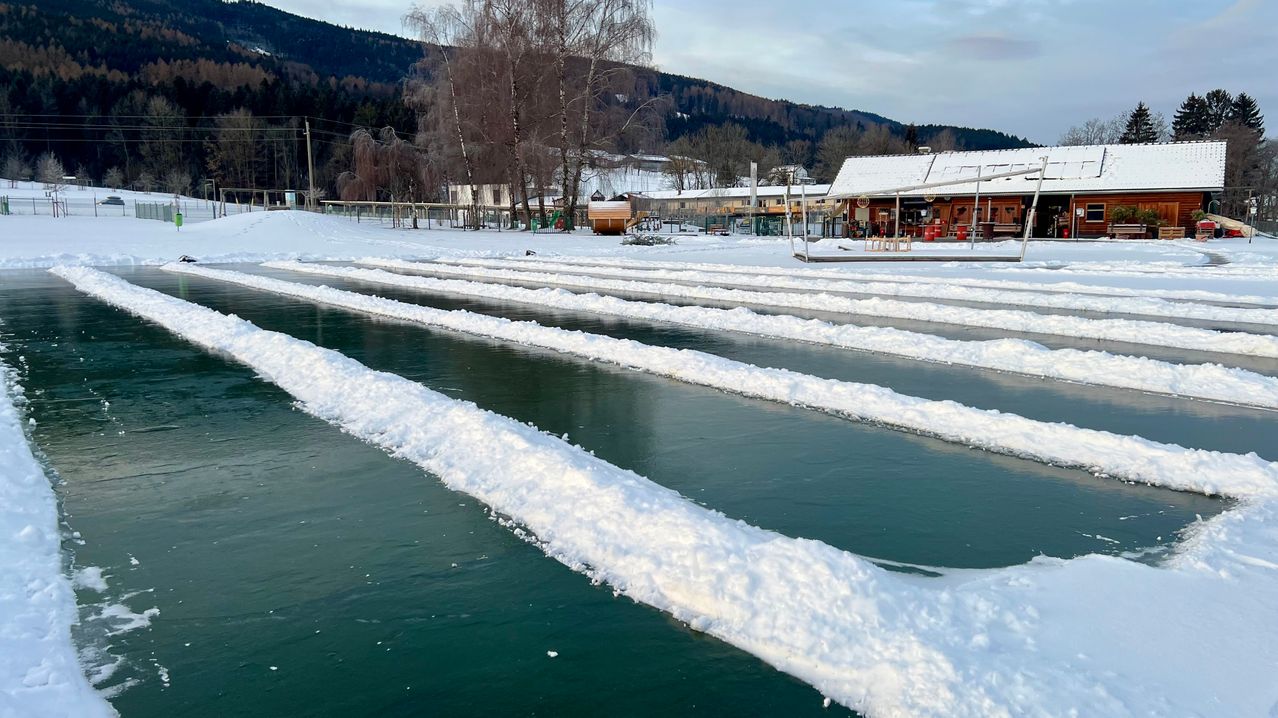 Eine verschneite Landschaft mit einem Gebäude, Bergen im Hintergrund und einem gefrorenen Fluss mit Schneehaufen an der Seite.