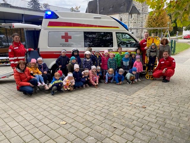 Eine Gruppe von Kindern und Erwachsenen, einige in Winterkleidung, posiert für ein Foto vor einem Krankenwagen. Der Krankenwagen hat ein rotes Kreuz an seiner Seite.
