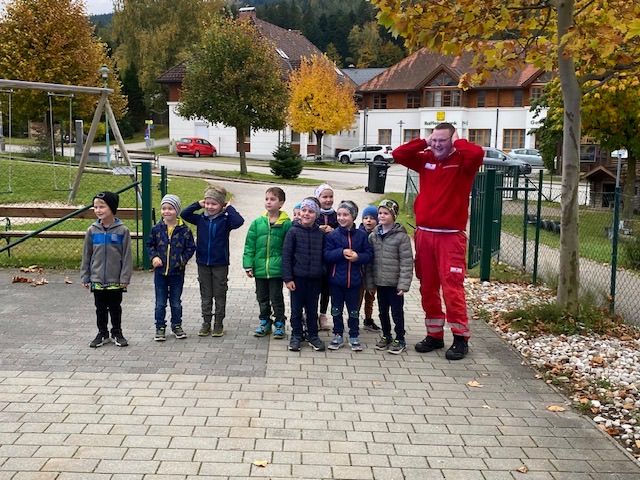 Eine Gruppe von Kindern und ein Mann in einem roten Anzug posieren für ein Foto in einem Park mit Bäumen und Gebäuden im Hintergrund.