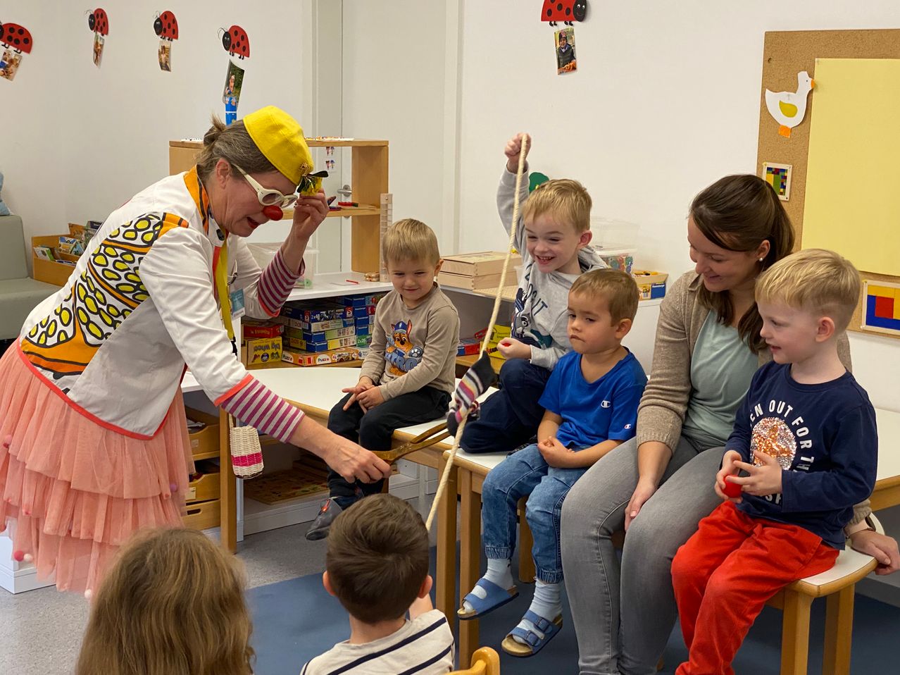 Ein Clown unterhält eine Gruppe von Kindern und Eltern in einem Spielzimmer. Der Clown hält eine Schnur und fesselt die Kinder, während sie auf Stühlen sitzen und aufmerksam zusehen.