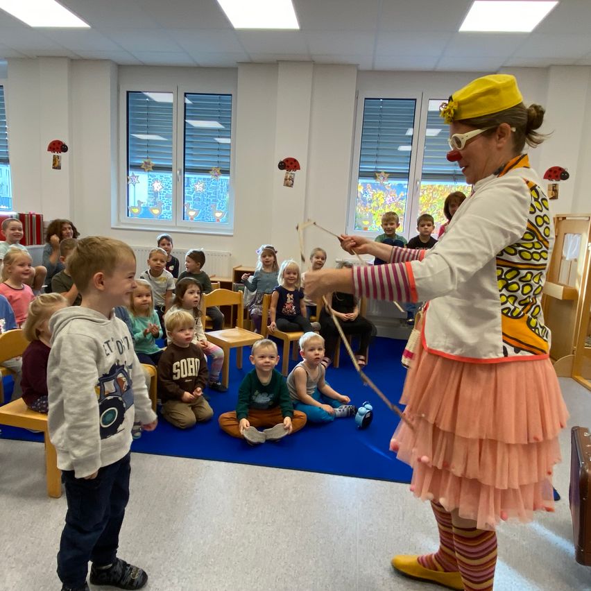 Ein Clown in einem bunten Kostüm tritt für eine Gruppe von Kindern im Klassenzimmer auf. Die Kinder sitzen auf dem Boden und beobachten den Clown.