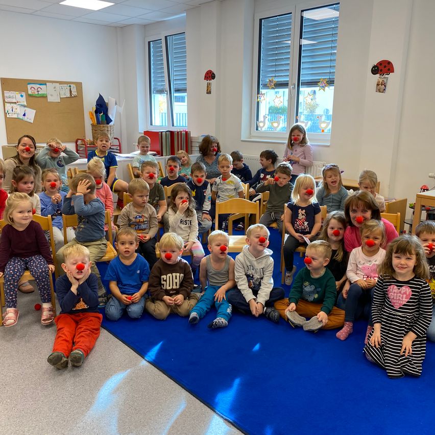 Ein Klassenzimmer voller Kinder mit roten Clownsnasen, die auf Stühlen auf einem blauen Teppich sitzen.