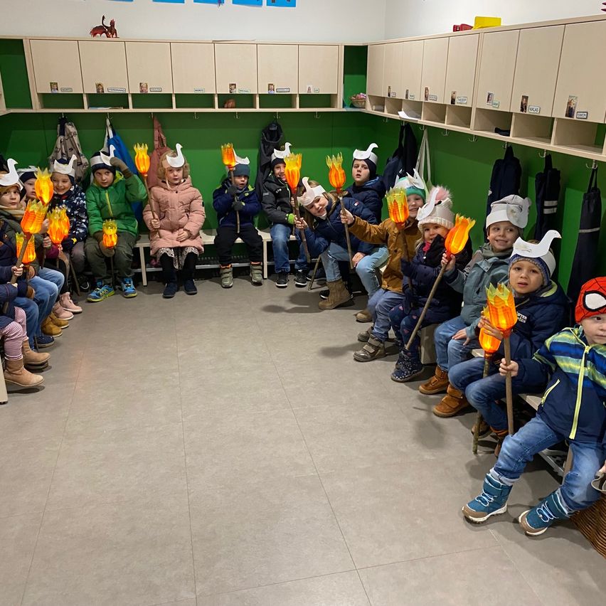 Eine Gruppe von Kindern in Winterkleidung und Mützen sitzt im Kreis und hält brennende Fackeln. Sie befinden sich in einem Raum mit grünen Wänden und Holzböden.