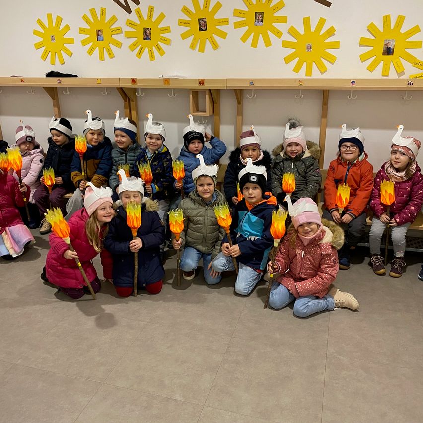 Eine Gruppe von Kindern, gekleidet in Winterkleidung, posiert für ein Foto. Sie halten Fackeln mit Flammen oben und es gibt sonenförmige Dekorationen an der Wand hinter ihnen.