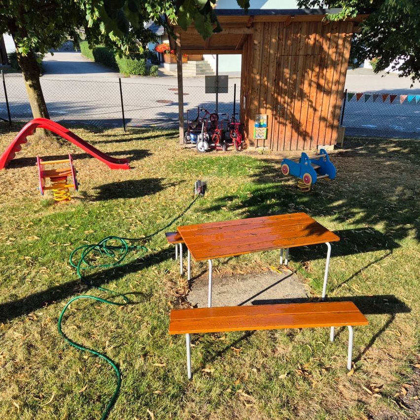 Ein Spielplatz mit einer Holzbank, einem Tisch und einer Rutsche. Ein kleines Holzhaus mit Spielzeug in der Nähe.