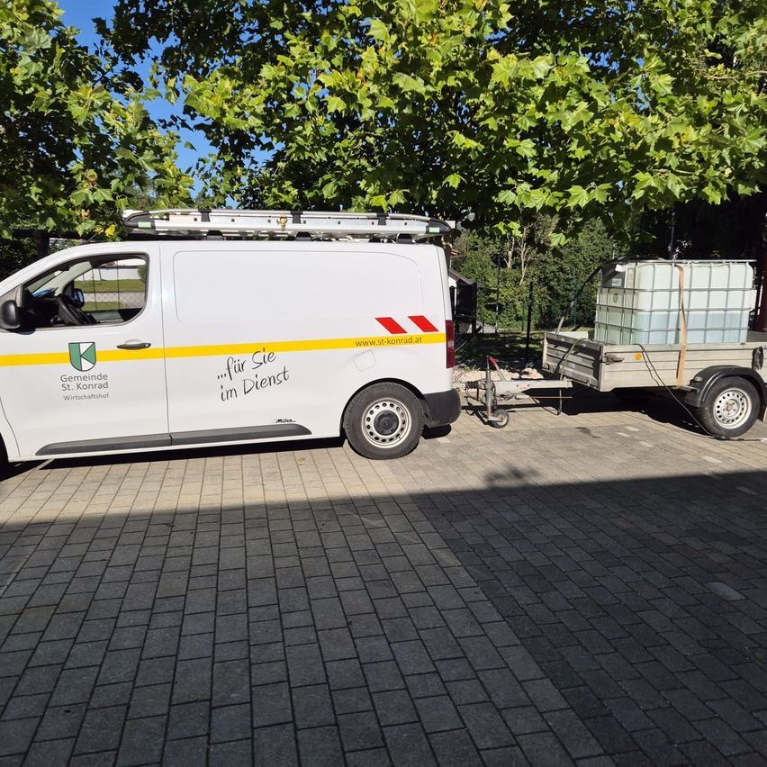 Ein weißer Lieferwagen mit gelbem Streifen und den Worten 'für Sie im Dienst' auf einer gepflasterten Einfahrt mit angehängtem Anhänger.