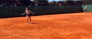 Eine Frau spielt Tennis auf einem Sandplatz bei klarem Himmel. Sie hält einen Tennisschläger und ist bereit, den Ball zu schlagen. Eine andere Person steht in der Ferne.