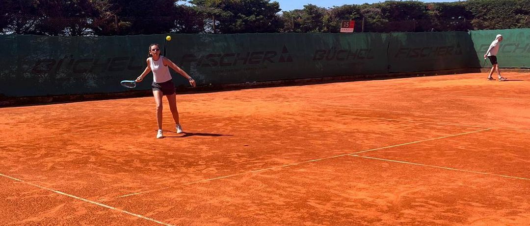 Eine Frau spielt Tennis auf einem Sandplatz bei klarem Himmel. Sie hält einen Tennisschläger und ist bereit, den Ball zu schlagen. Eine andere Person steht in der Ferne.