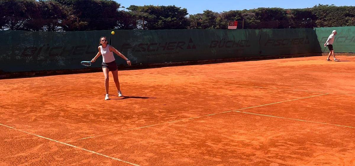 Eine Frau spielt Tennis auf einem Sandplatz bei klarem Himmel. Sie hält einen Tennisschläger und ist bereit, den Ball zu schlagen. Eine andere Person steht in der Ferne.