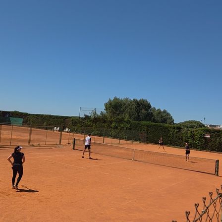An einem sonnigen Tag spielen vier Personen Tennis auf einem Freiplatz. Eine Person steht links, und drei sind auf der rechten Seite. Das Netz teilt den Platz. Bäume und Büsche umgeben das Gebiet.
