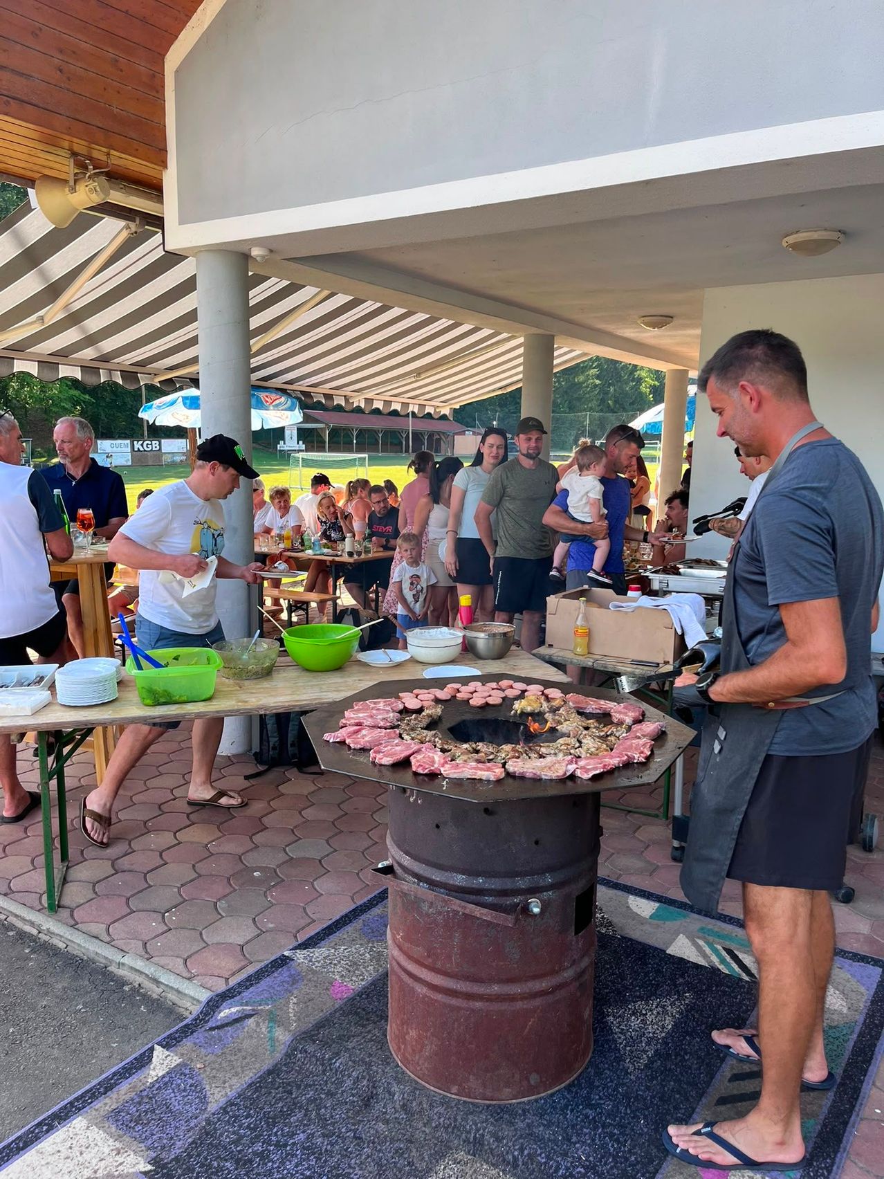 A man cooks meat on a grill while others wait in line at an outdoor restaurant.