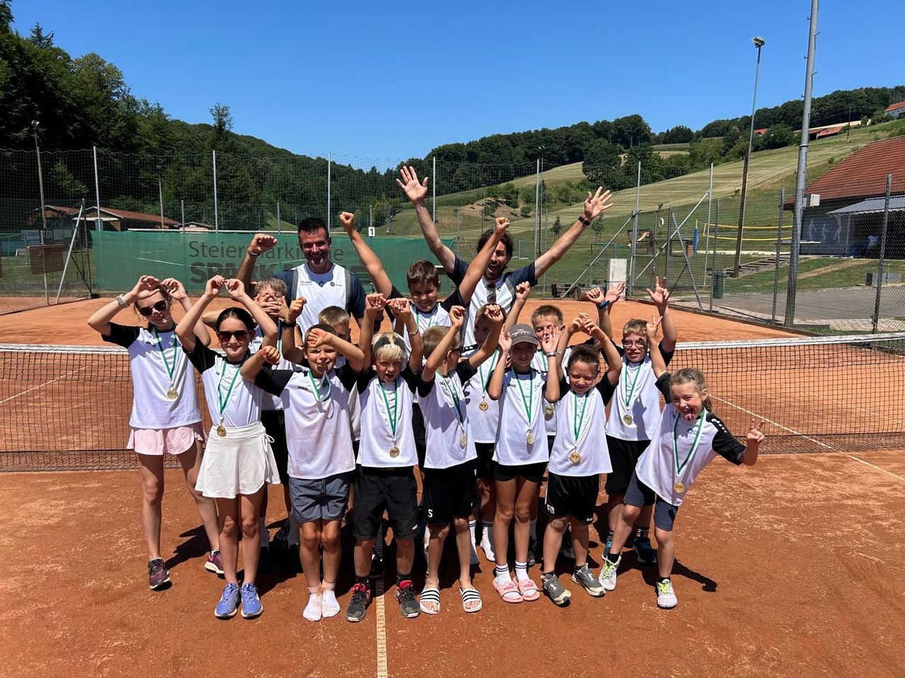 A group of young tennis players with medals around their necks pose for a photo on a tennis court.