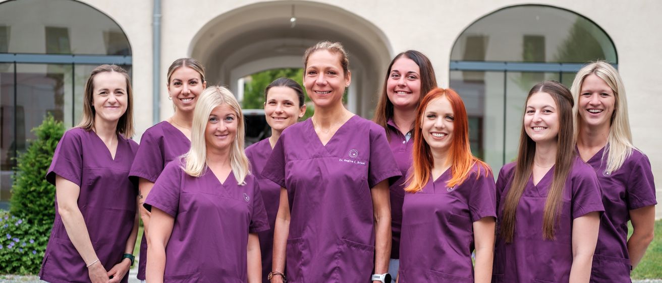 Eine Gruppe lächelnder Frauen, angezogen in passenden lila Kitteln und weißen Hosen, steht zusammen vor einem Gebäude.