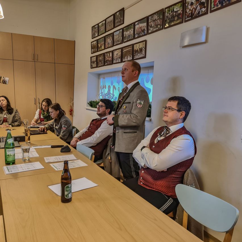 Eine Gruppe von Menschen in einem Besprechungsraum, wobei ein Mann steht und spricht. Es gibt Schreibtische mit Papieren und Flaschen, und gerahmte Bilder an der Wand.