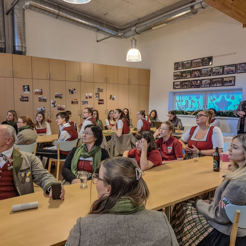 Eine Gruppe von Menschen sitzt in einem Raum mit Tischen und Stühlen. Einige tragen rote Westen. Flaschen und Gläser stehen auf den Tischen. Bilderrahmen sind an der Wand montiert.