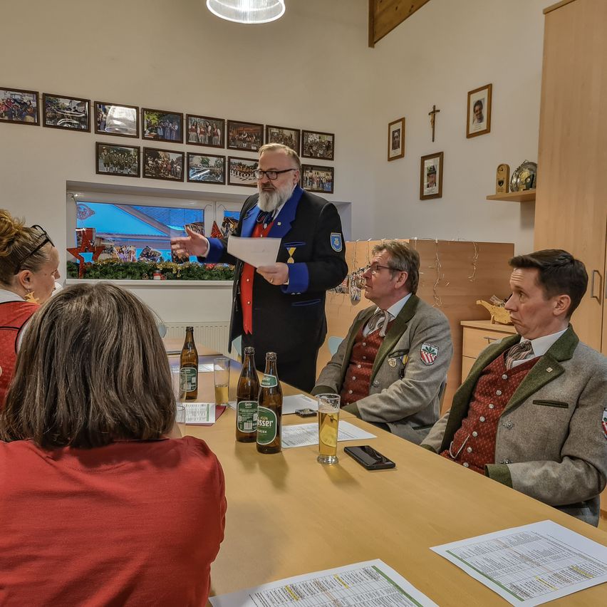 Ein Mann mit einer roten Schleife steht an einem Tisch mit vier sitzenden Personen. Sie alle sind in traditioneller Kleidung gekleidet. Ein Glas Bier, zwei Flaschen und ein Handy stehen auf dem Tisch. Über ihnen befinden sich Bilderrahmen, und ein Fenster zeigt eine festliche Außenszene.