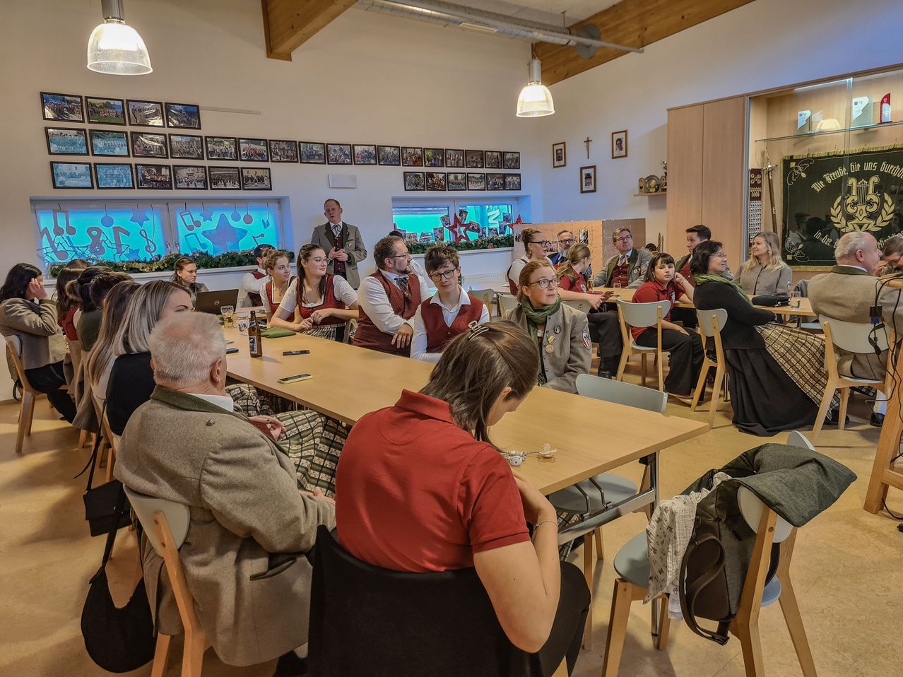 Eine Gruppe von Menschen sitzt um einen Tisch in einem Raum mit Holzwänden und Decken. Einige Personen tragen Brillen und Jacken. Flaschen und ein Laptop sind auf dem Tisch. Eine Person sitzt auf einem Stuhl und blickt nach unten.