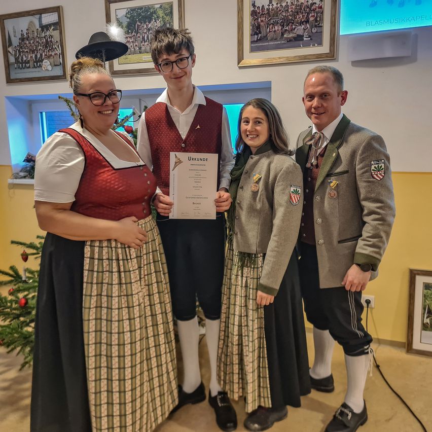 Vier Personen stehen in einem Raum, lächeln und posieren für ein Foto. Ein Mann hält eine Urkunde. Zwei Frauen tragen traditionelle Kleidung. Ein Weihnachtsbaum und gerahmte Bilder sind im Hintergrund.