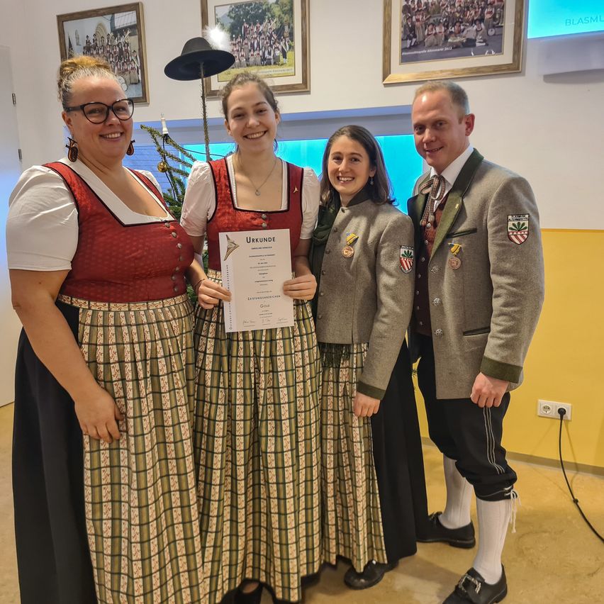 Vier Personen posieren für ein Foto, zwei Frauen in traditioneller Kleidung, eine hält ein Zertifikat, und ein Mann in formeller Kleidung, mit einem Weihnachtsbaum und gerahmten Fotos im Hintergrund.