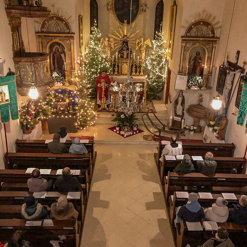 Ein Innenraum einer Kirche mit einem Priester am Altar. Der Altar ist mit einem Weihnachtsbaum und Kerzen geschmückt. Die Kirchenbänke sind mit Menschen gefüllt. Die Kirchenwände sind mit religiösen Bildern und Statuen verziert.