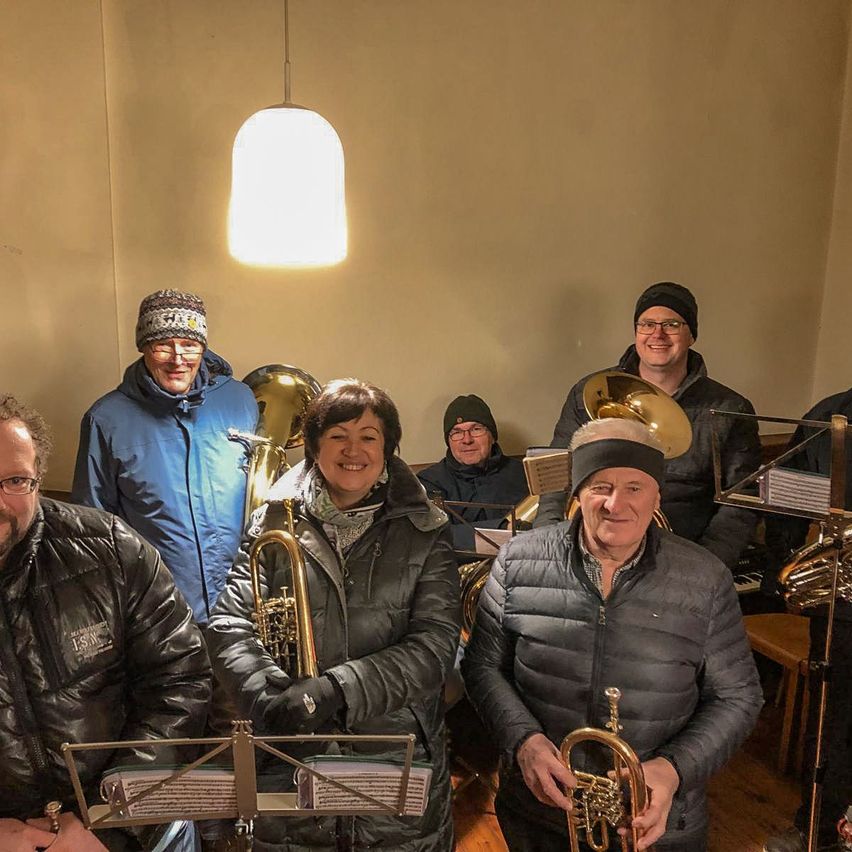 Eine Gruppe von Musikern versammelt sich in einem Raum, alle warm gekleidet. Sie halten Blechblasinstrumente, darunter Tubas und Trompeten. Eine Frau in der Mitte lächelt, und der Raum wird von einer hängenden Lampe beleuchtet.