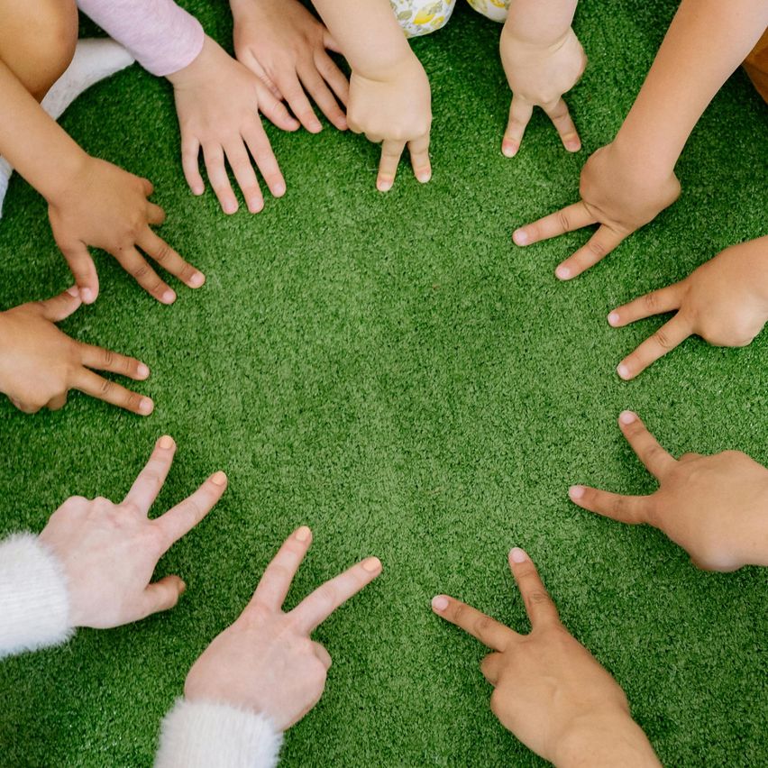 Eine Gruppe von Kindern sitzt im Kreis auf grünem Teppich und bildet mit ihren Fingern ein V-Zeichen. Sie alle schauen nach vorne und tragen Freizeitkleidung.