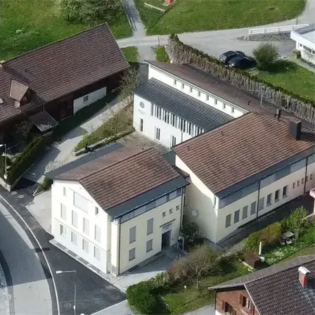 Aerial view of a rural area with multiple buildings. A large brown house and two smaller white buildings with brown roofs. Solar panels on one of the white buildings. A road with a parked car.