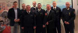 Mehrere Männer in Uniformen stehen zusammen und lächeln für ein Foto. Sie stehen vor einem großen Banner mit deutschem Text und einer Illustration eines Feuerwehrwagens.